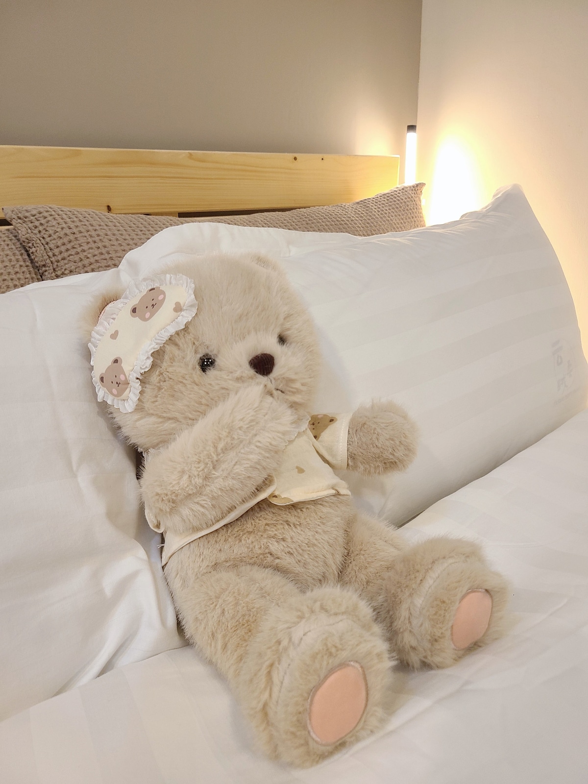 A plush teddy bear is positioned on a neatly arranged bed, featuring white linens with subtle stripes. The bear is dressed in a light-colored outfit and holds a playful expression, nestled against soft pillows in a muted color palette.