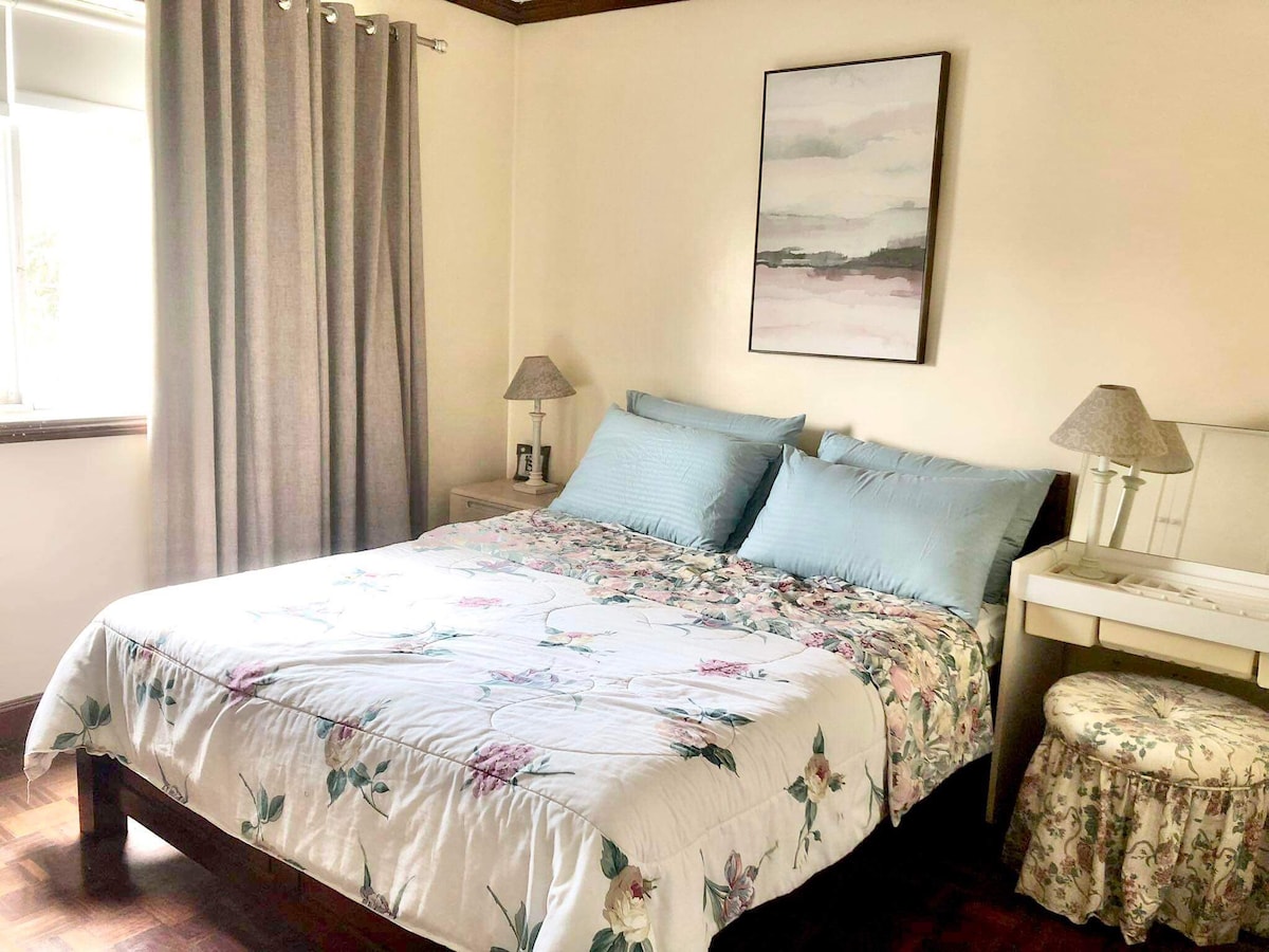 A spacious bedroom is featured, centered around a large bed dressed with a floral quilt. Soft blue pillows accent the bedding. Natural light enters through a window dressed with light curtains, while a small bedside table and lamp complement the cozy atmosphere.