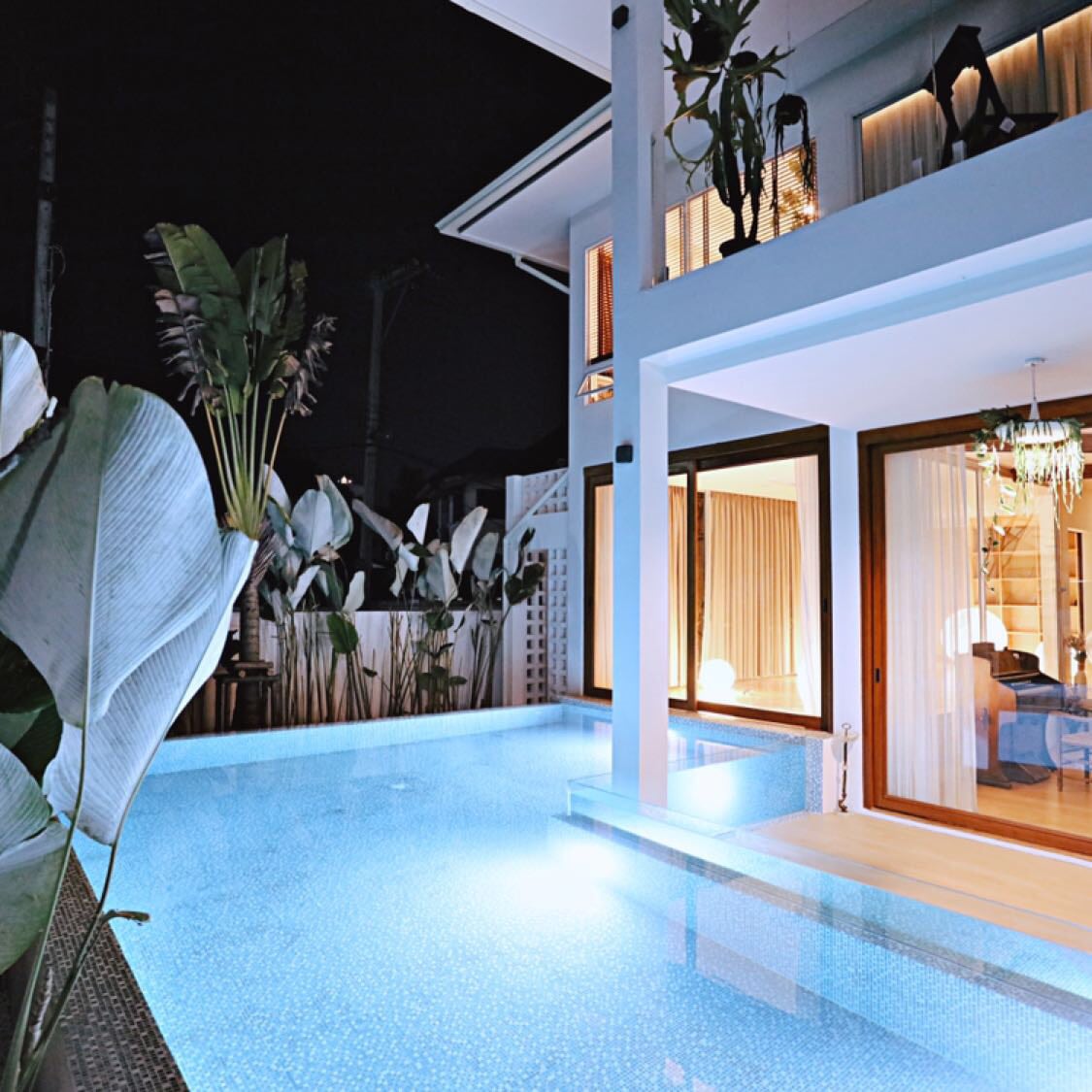 A serene outdoor area is highlighted, featuring a well-lit swimming pool with turquoise water reflecting the night sky. Large plants frame the space, while expansive glass doors lead into the interior, allowing for a seamless transition between the indoor and outdoor environments.