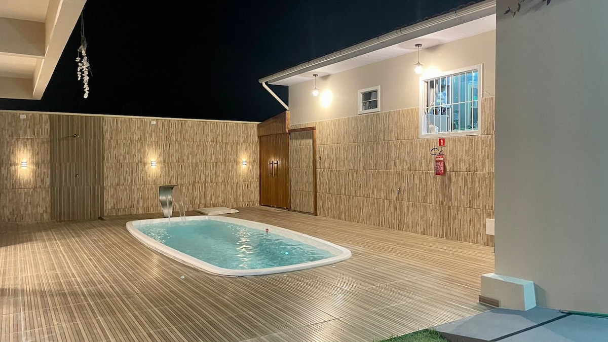 A private pool area is illuminated by soft overhead lights, showcasing a rectangular swimming pool surrounded by wooden decking. The surrounding walls are lined with natural materials, creating a serene environment. Doors leading to adjacent facilities are visible in the background.