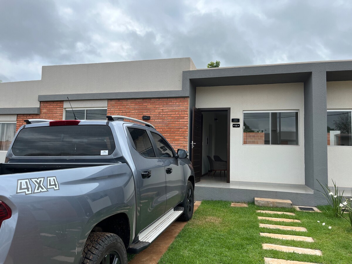 A modern exterior is presented, featuring a sleek gray vehicle parked on a pathway leading to the entrance. The building's façade combines light gray and brick materials, with two large windows flanking the front door, which opens to a welcoming interior.