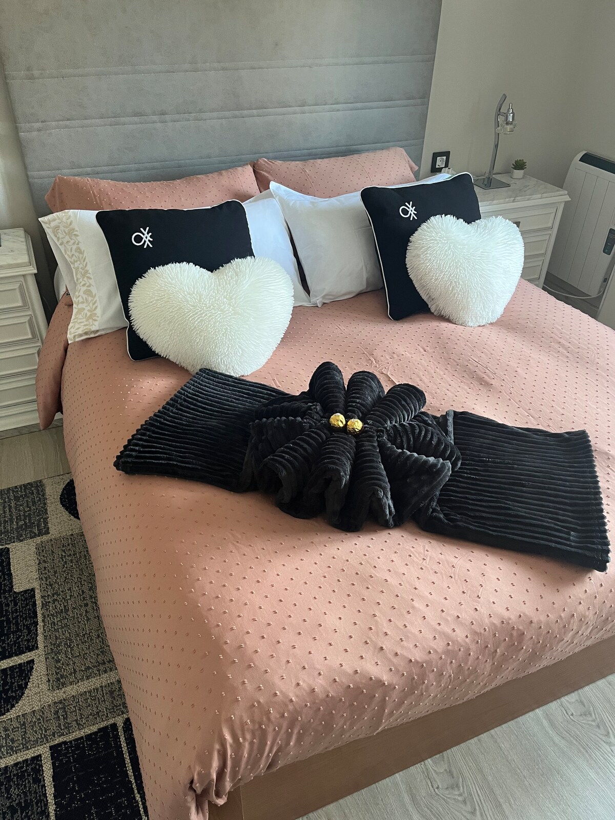 A spacious bed is presented with a soft coral-colored coverlet and layered with white and black decorative pillows in heart shapes. A decorative towel arrangement is placed at the foot of the bed, adding a touch of elegance to the room.