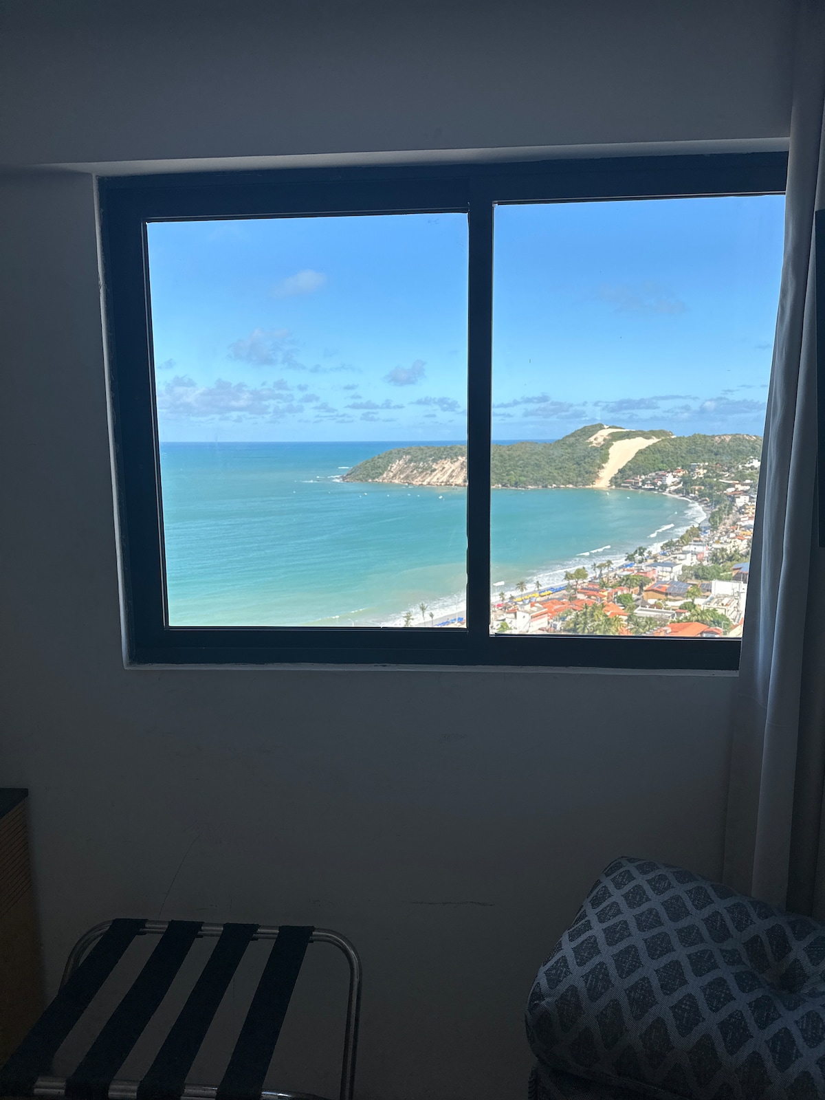 A large window frames a stunning view of the ocean and coastline, showcasing clear blue waters and a sandy beach. The scenic landscape includes hills and a portion of the city below, under a bright sky with scattered clouds.