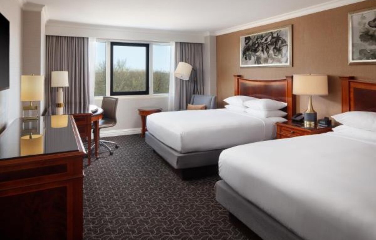 A well-appointed guest room features two spacious beds flanked by nightstands and lamps. A work desk is positioned near a window, offering views of greenery outside. Neutral tones and soft textures create a serene environment, complemented by artworks on the walls.