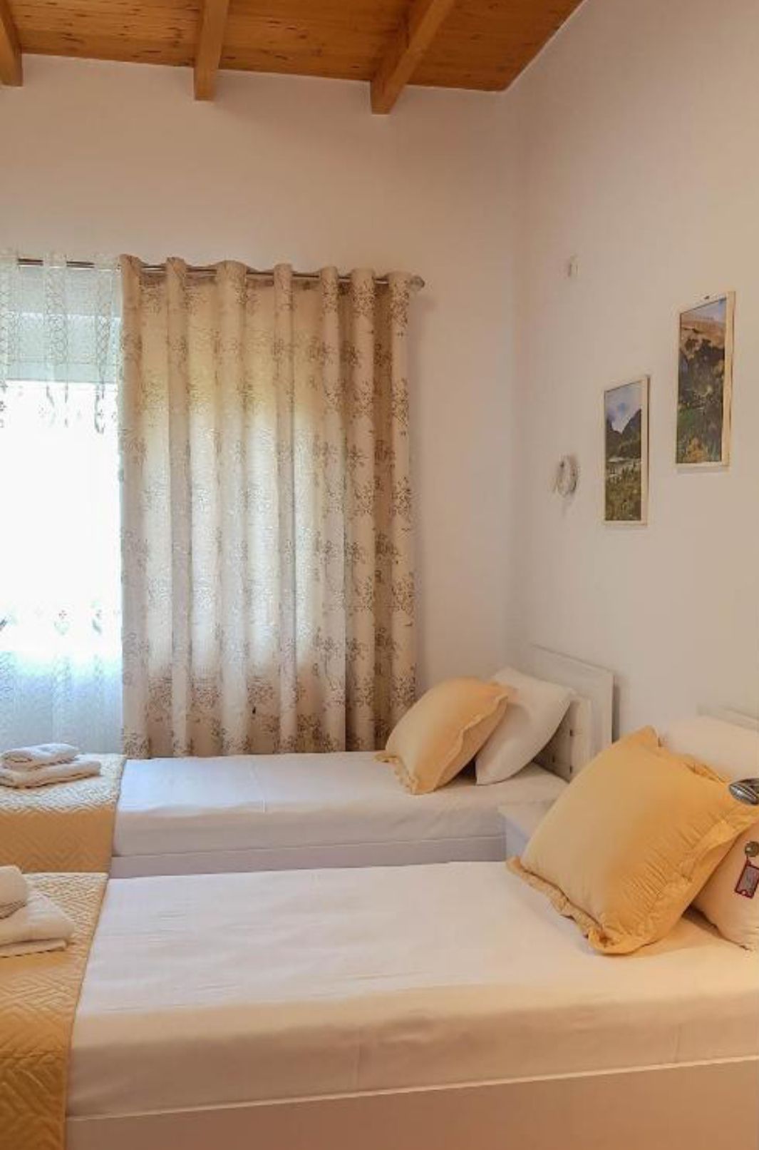 A serene sleeping area features two single beds dressed in white linens and yellow accent pillows. Natural light filters through a window covered with sheer curtains, illuminating the space. Light-colored walls are complemented by soft, patterned drapes and framed wall art.