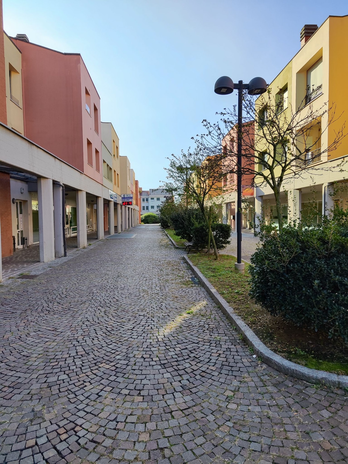 A cobblestone pathway winds through a colorful residential area, flanked by buildings in soft shades of pink and yellow. Small shrubs are seen along the sides, while trees provide gentle shade. Street lamps stand tall, offering illumination for evening strolls.