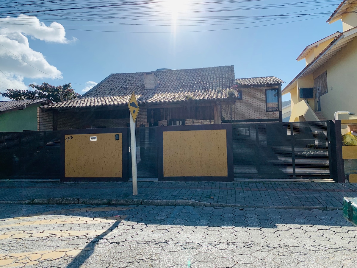 The exterior of the chalet showcases a two-story structure with rustic brickwork and wooden beams. A gated entrance is visible, flanked by a stone-paved road. Natural sunlight illuminates the area, highlighting the traditional roof and surrounding greenery.