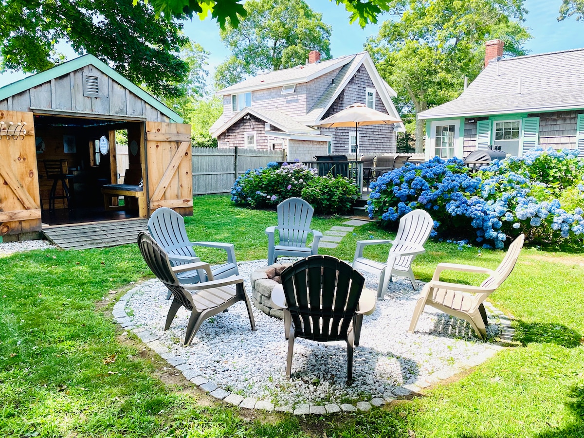 An outdoor seating area features a circular arrangement of Adirondack chairs around a fire pit, set on a pebbled surface. Bright blue hydrangeas bloom nearby, complementing the lush green lawn and the wooden shed in the background under a clear sky.