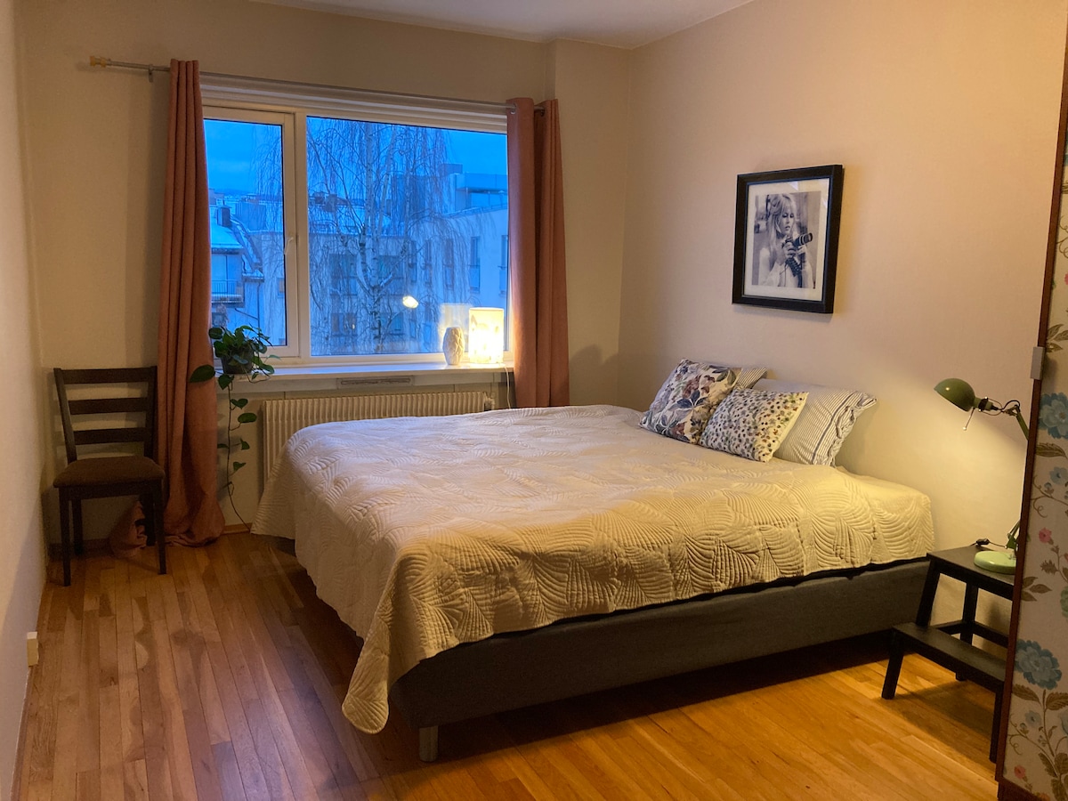 A serene bedroom is presented with a spacious bed featuring a light-colored quilt. A window allows natural light to fill the room, offering a view of the surrounding area. A small chair and a bedside table with a lamp add functional touches to the space.