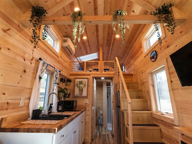 The interior of the tiny home showcases natural wood paneling, contributing to a warm ambiance. A staircase leads to the upper level, while large windows allow ample natural light. Plants hang from the ceiling, and a cozy kitchen area is visible, equipped with modern appliances.