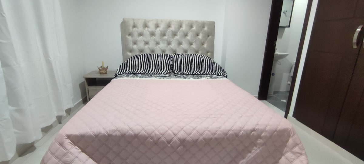 A spacious bed with a tufted headboard is presented, adorned with striped pillows and a soft pink quilt. Natural light filters through the sheer curtains, creating a calm and inviting atmosphere. An elegant bedside table sits adjacent, featuring a decorative item.