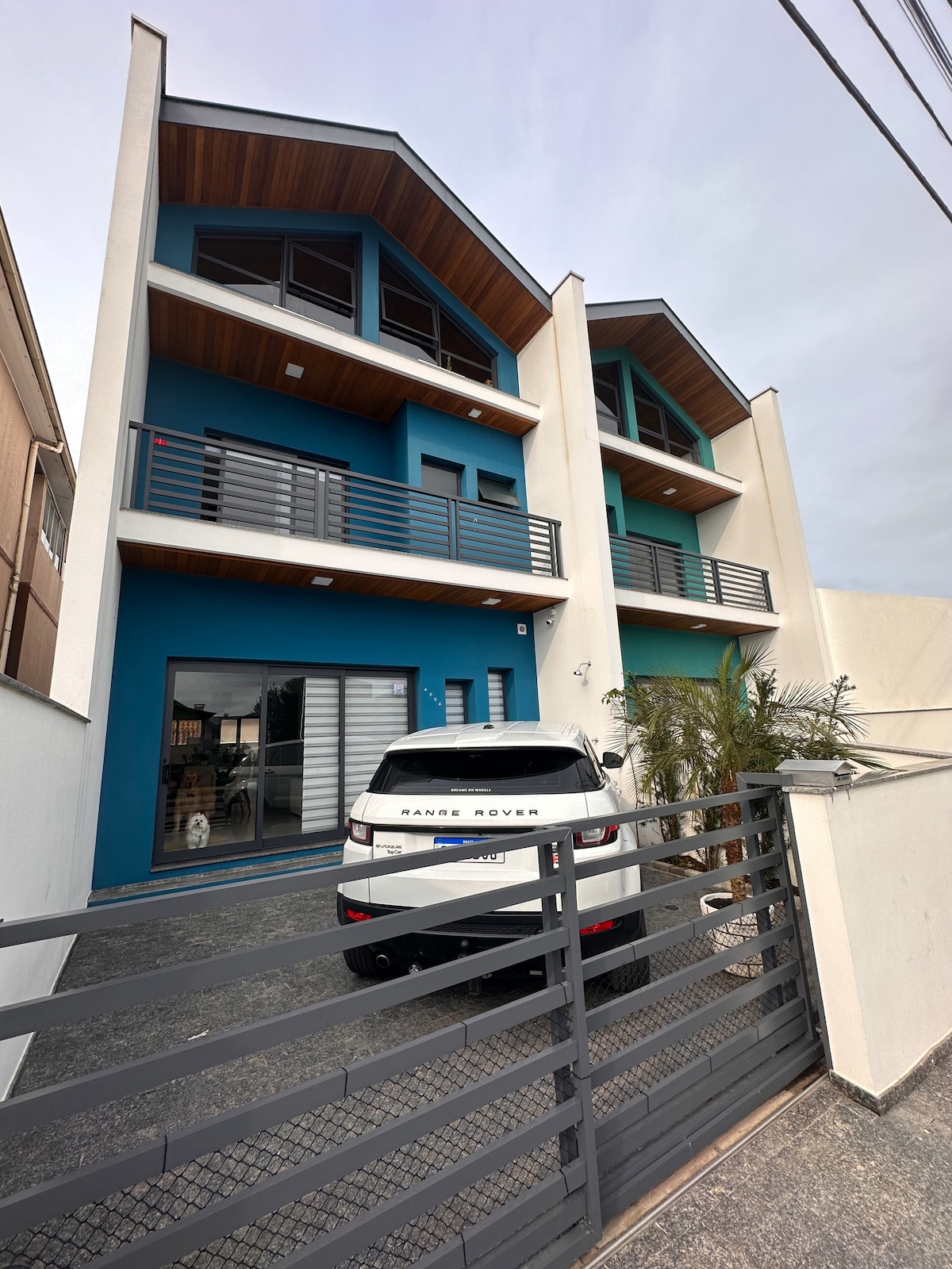 A modern triplex structure is presented in blue and white tones, featuring a clean exterior with balconies and large windows. A paved driveway is visible, accommodating a vehicle. Lush greenery adds to the welcoming entry, creating an inviting atmosphere for guests.