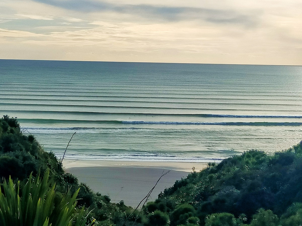 A serene coastal view is presented, featuring gently rolling waves that form rhythmic lines across the horizon. A sandy beach is visible below, flanked by lush greenery that leads to a tranquil ocean reflecting soft hues of blue and green under a cloudy sky.