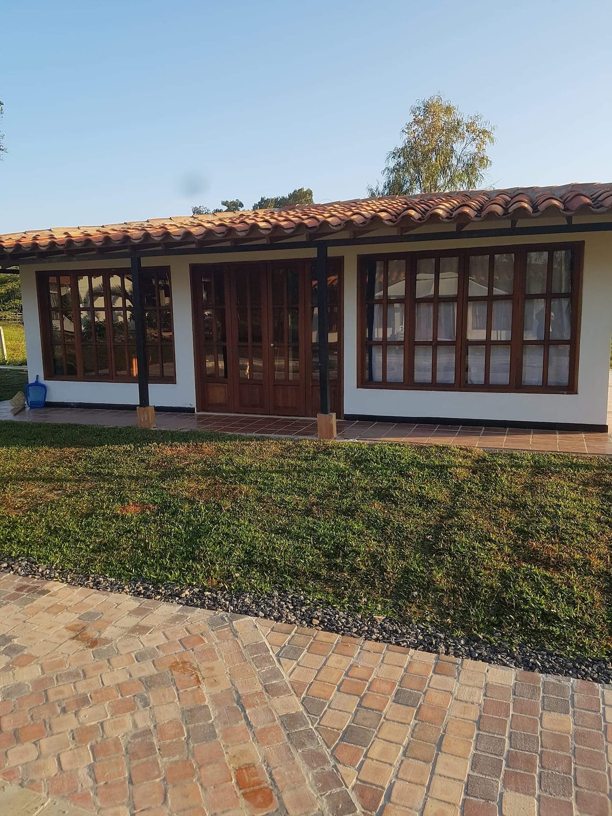 A charming cabin features large windows framed in wood, allowing natural light to fill the interior. The exterior is complemented by a tiled roof and lush greenery surrounding the entrance. A paved pathway leads to the front door, enhancing accessibility.