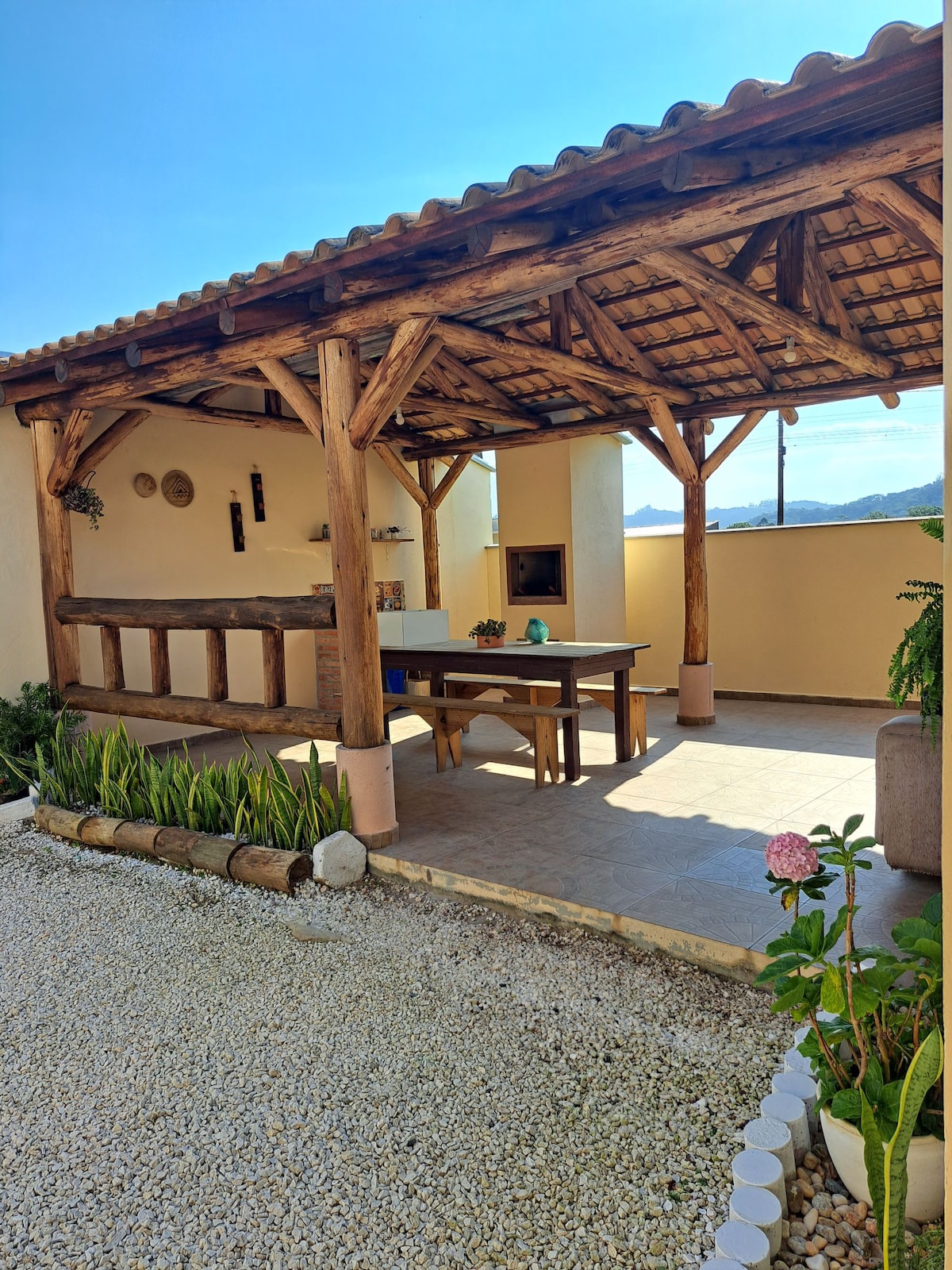 An outdoor area is shown, featuring a wooden roof supported by beams. A large table is centered beneath the shelter, accompanied by seating. A barbecue grill is situated against the back wall, while plants and decorative stones line the perimeter, enhancing the inviting space.