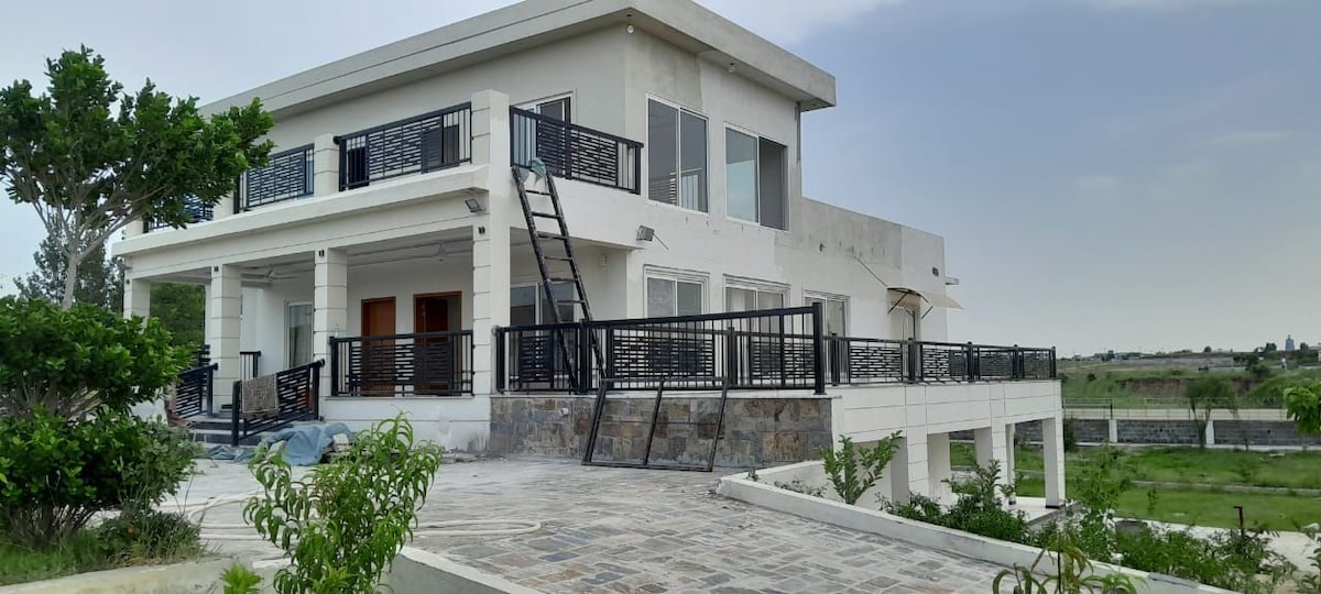 A modern white structure features large windows and multiple balconies, providing expansive views of the surrounding landscape. A stone pathway leads to the entryway, flanked by greenery. The outdoor area includes a spacious patio made of stone, offering ample space for relaxation.