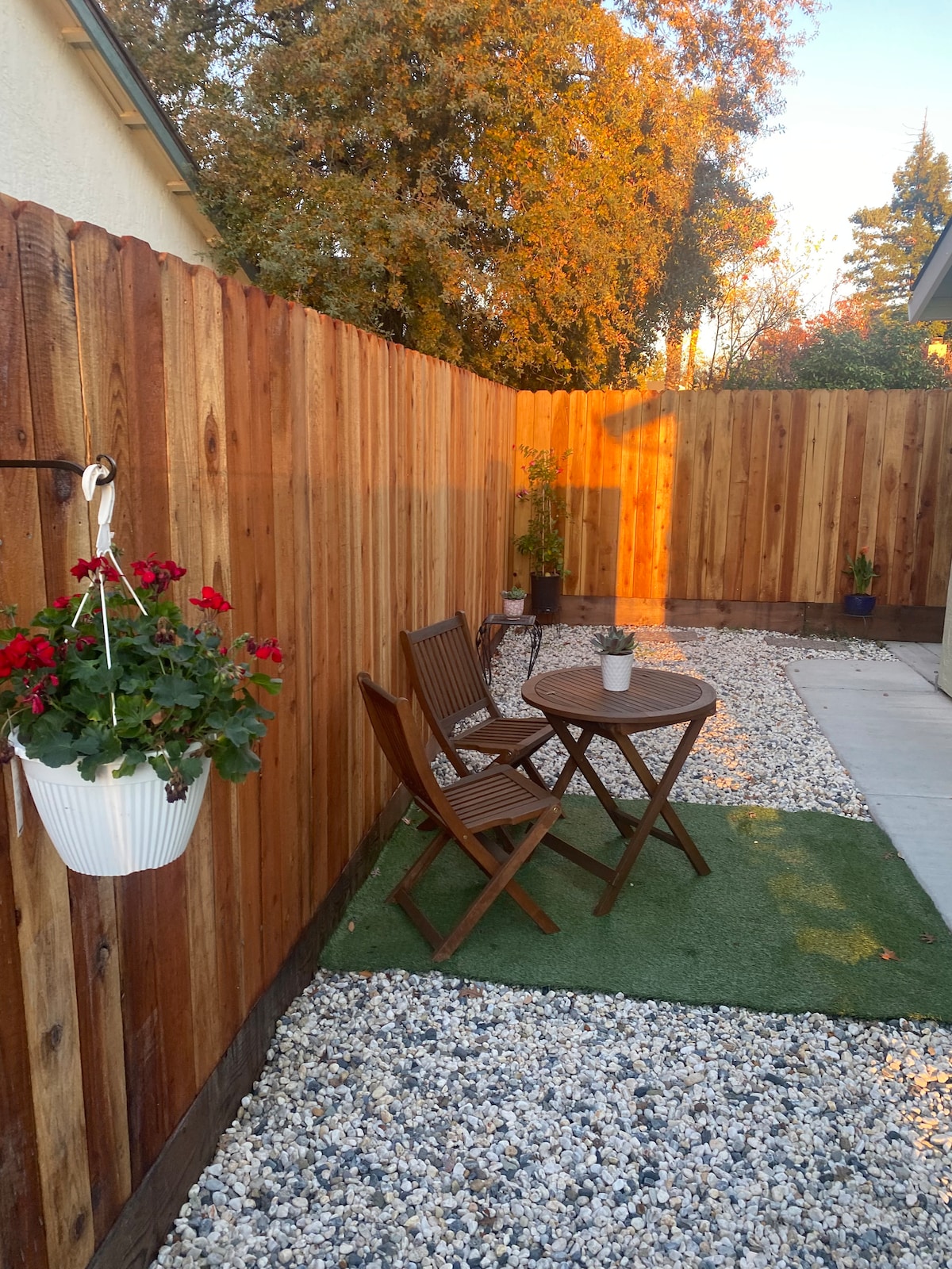 An outdoor space is enclosed by a wooden fence, featuring a small round table and two wooden chairs on a gravel surface. A hanging planter with red flowers adds a touch of color to the setting. Green grass is placed beneath the furniture, enhancing the cozy ambiance.
