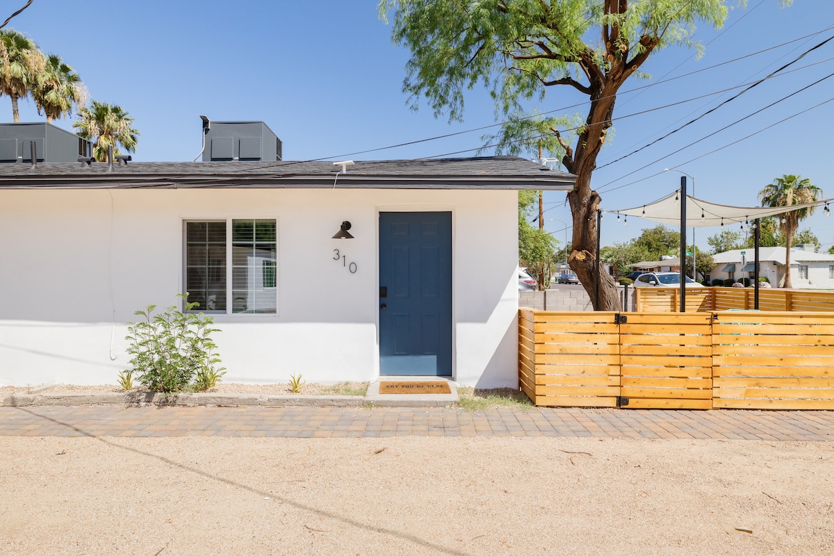 The front entrance features a bright blue door surrounded by a clean white exterior. A linear pathway leads up to the door, flanked by minimal landscaping and a wooden privacy fence. An inviting atmosphere is created by the nearby trees and clear sky.