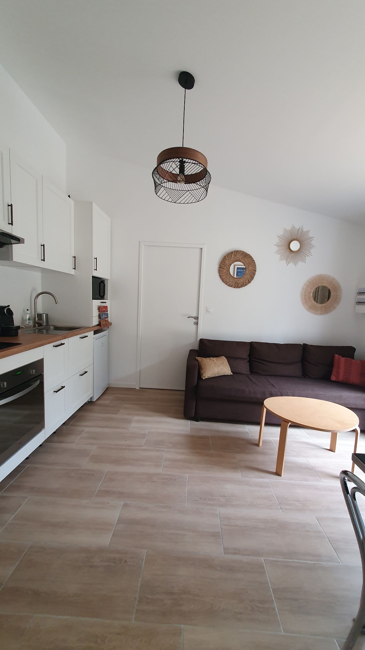 The cozy living area features a compact kitchen equipped with modern appliances and white cabinetry. A comfortable dark sofa with decorative cushions faces a round wooden coffee table. The walls are adorned with decorative mirrors, enhancing the natural light that fills the space.