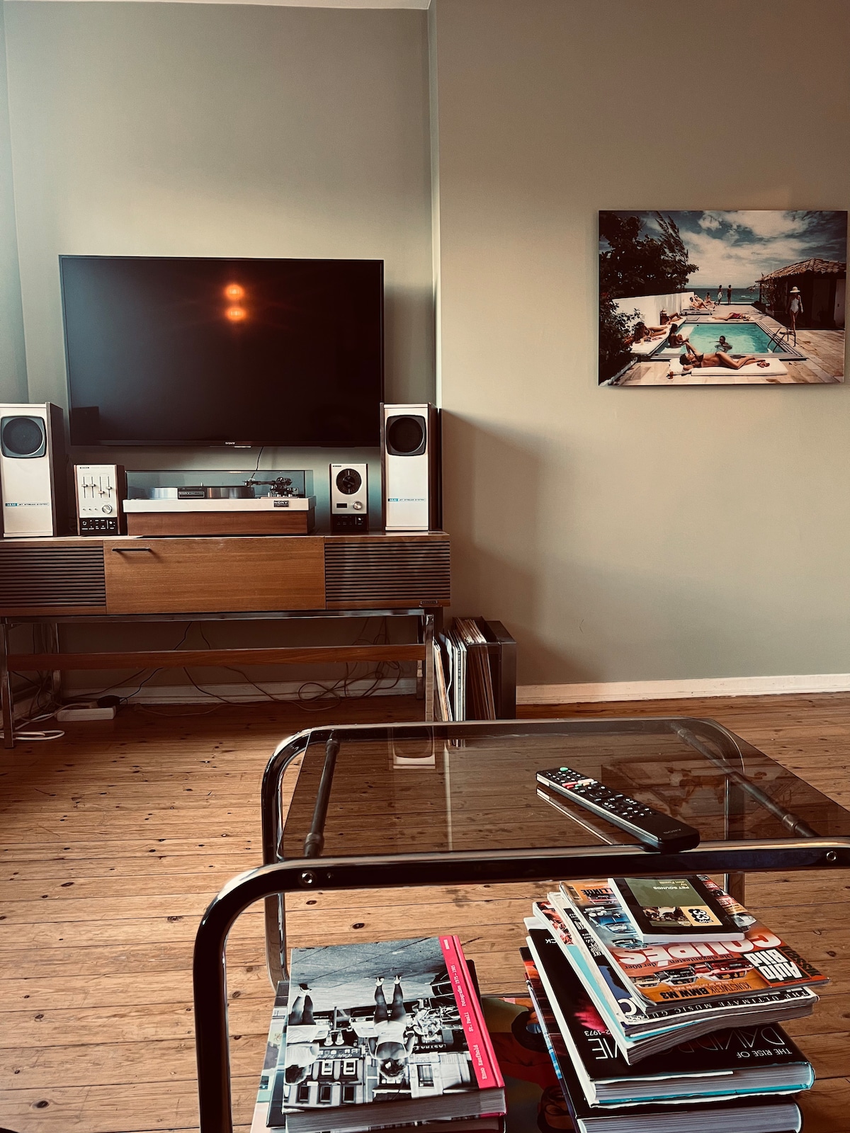 A living area features a flat-screen television mounted above a wooden media console, accompanied by speakers on either side. A glass coffee table displays magazines and a remote control. A vibrant artwork is mounted on the wall, depicting a poolside scene.