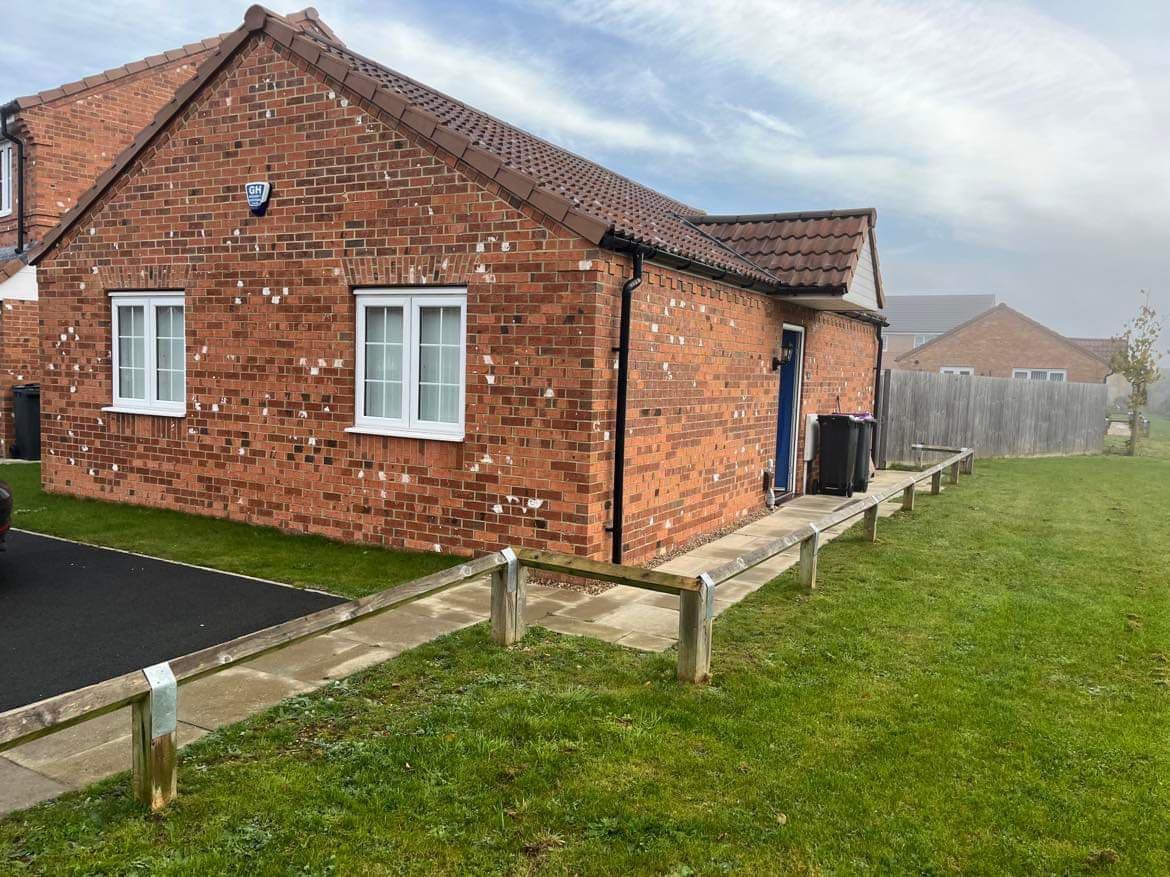 A single-story bungalow is presented with a brick exterior and a tiled roof. The property includes a neatly landscaped front yard with grass and a pathway leading to the entrance. Nearby parking spaces are visible, along with a wooden fence in the background.