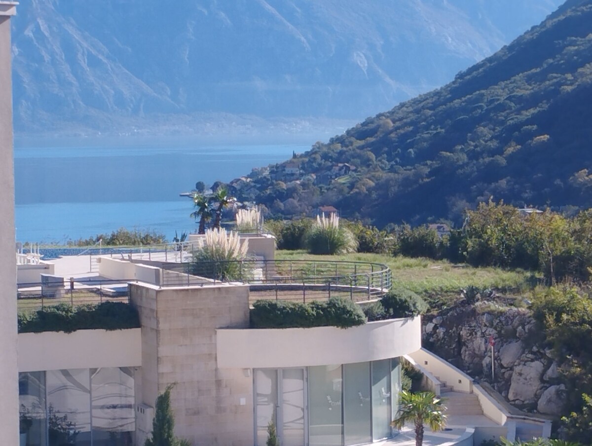 A spacious terrace features lush greenery and decorative plants with panoramic views of Boka Bay. The nearby mountains rise majestically in the background, creating a serene natural setting. Sunlight reflects off the calm water, enhancing the peaceful atmosphere.