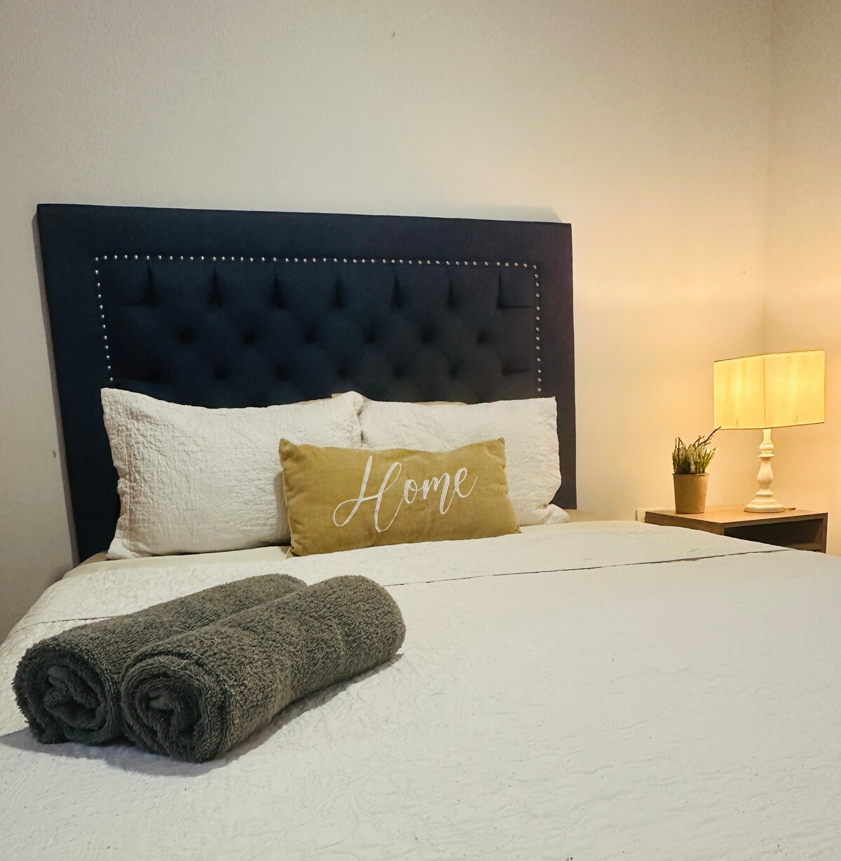 A spacious bed is adorned with a tufted navy headboard and crisp white linens. Two decorative pillows rest at the head, accompanied by a cozy throw blanket. A bedside table with a lamp and a small plant adds a touch of warmth to the restful space.