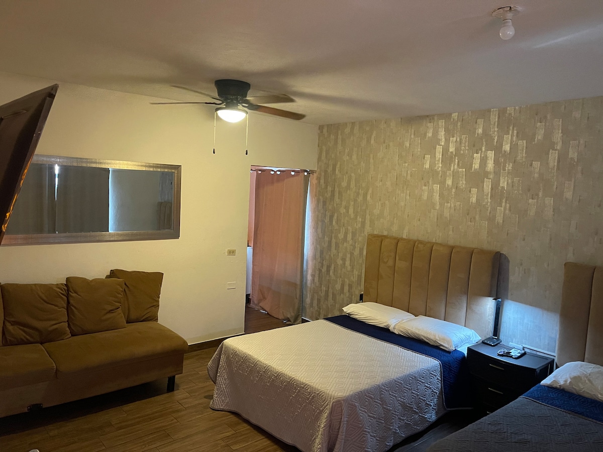 A spacious room is presented with a seating area featuring a brown couch. Two beds are visible, one covered with a blue blanket and the other with a white quilt. A ceiling fan and a mirror enhance the room's functionality.