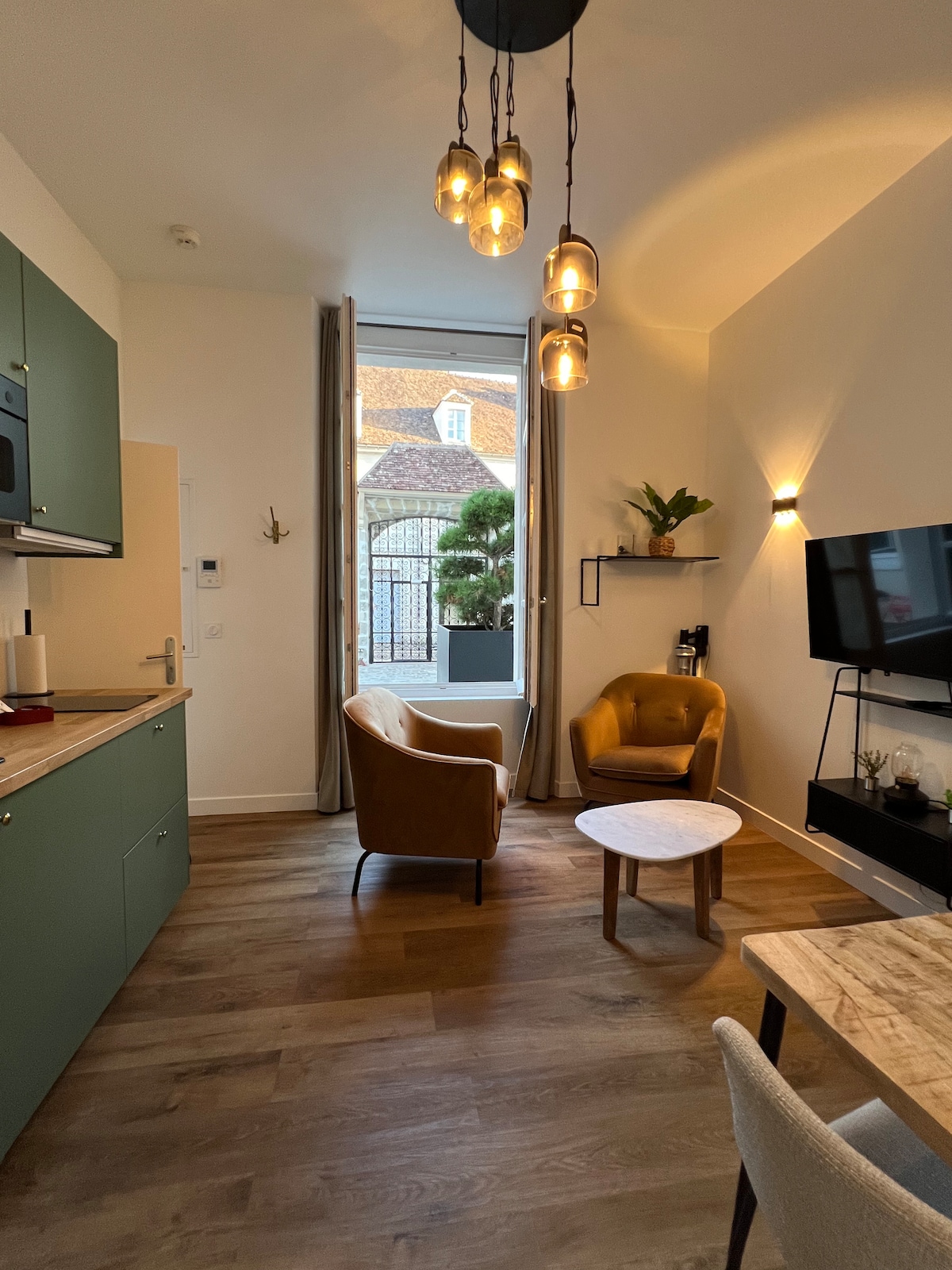A cozy living area features two comfortable chairs positioned beside a small round table. Natural light streams through a large window, highlighting the warm wood flooring and modern decor. A wall-mounted TV is visible, along with decorative shelves that add character to the space.