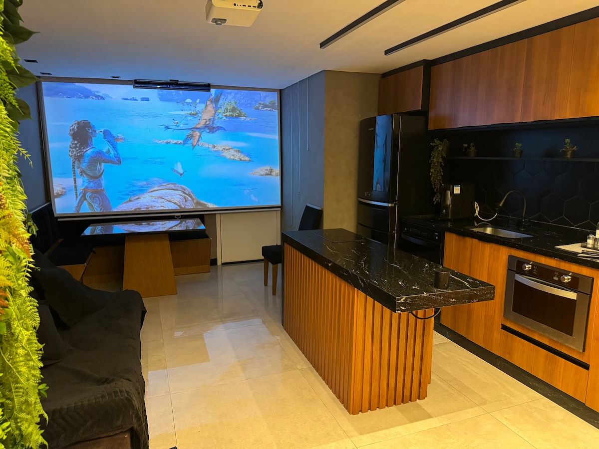 A home cinema space is showcased, featuring a large projector screen displaying a vibrant scene. The kitchen area includes modern appliances with a sleek black countertop and a wooden breakfast bar. A dark sofa and dining table complete the cozy, functional layout.