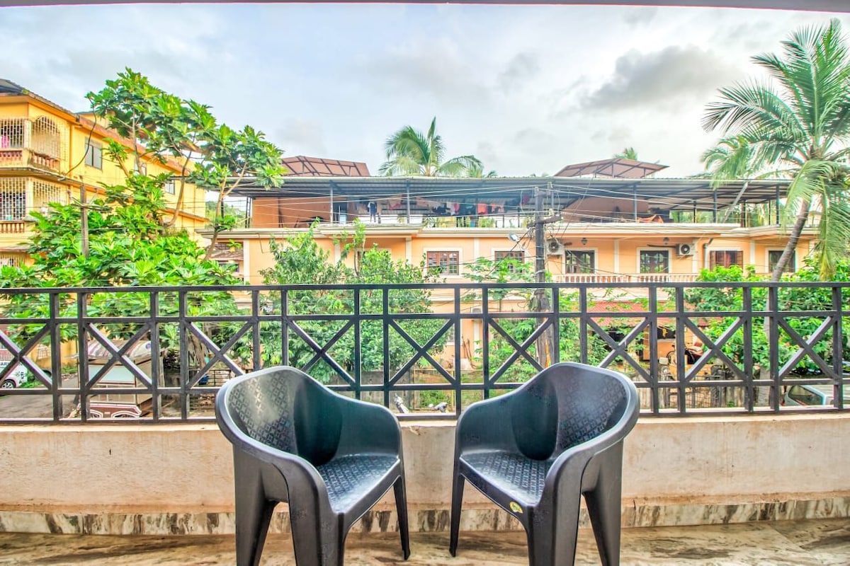 A balcony space is presented with two black chairs overlooking lush greenery. The view includes a mix of residential buildings with palm trees, adding a touch of nature to the area. The atmosphere appears calm and inviting.