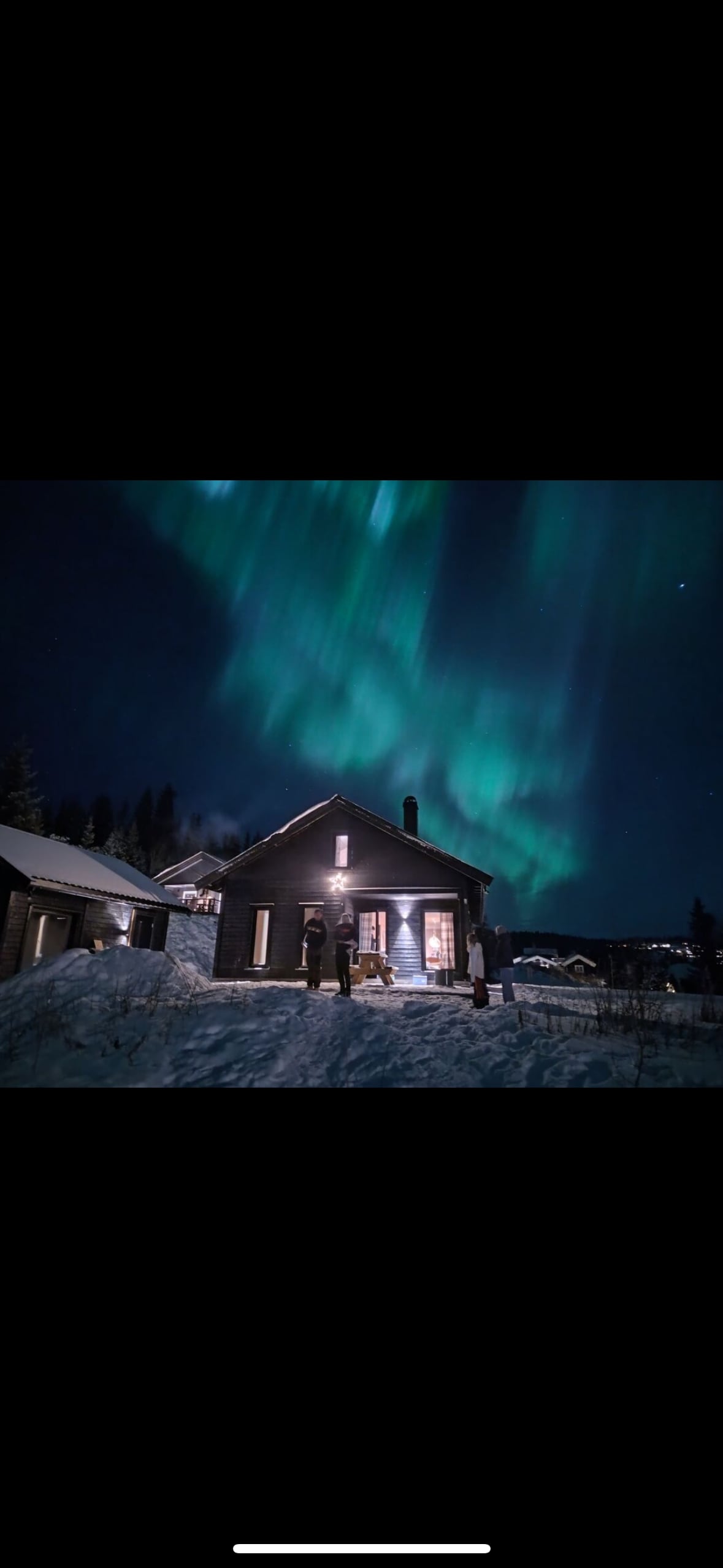 A charming cabin is illuminated under a stunning display of the northern lights. The structure features a warm glow from the windows, surrounded by snow-covered ground and distant hills, creating a serene winter landscape.