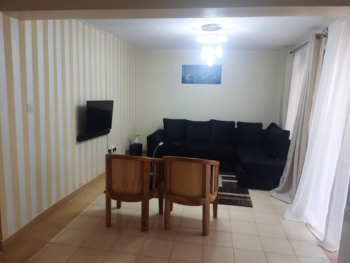 A living area features a comfortable black sectional couch positioned against a light wall. A flat-screen TV is mounted nearby. Two wooden chairs are arranged around a small table, with natural light illuminating the space through sheer curtains.