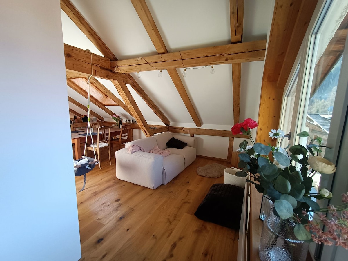 The open living area features exposed wooden beams across the ceiling, providing a warm feel. A light-colored sofa sits on the wooden floor, accompanied by a cozy rug. In the background, a dining table with chairs is visible, along with potted plants near a window.