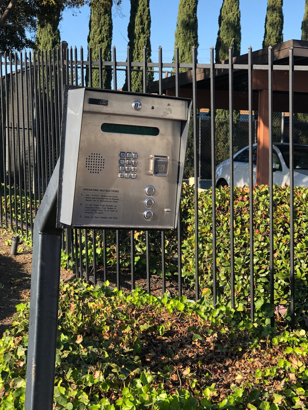A metallic intercom system is mounted on a curved stand, providing access to a gated entrance. Dense green foliage and a black fence surround the area, with tall coniferous trees visible in the background. A car is parked under a shaded structure nearby.