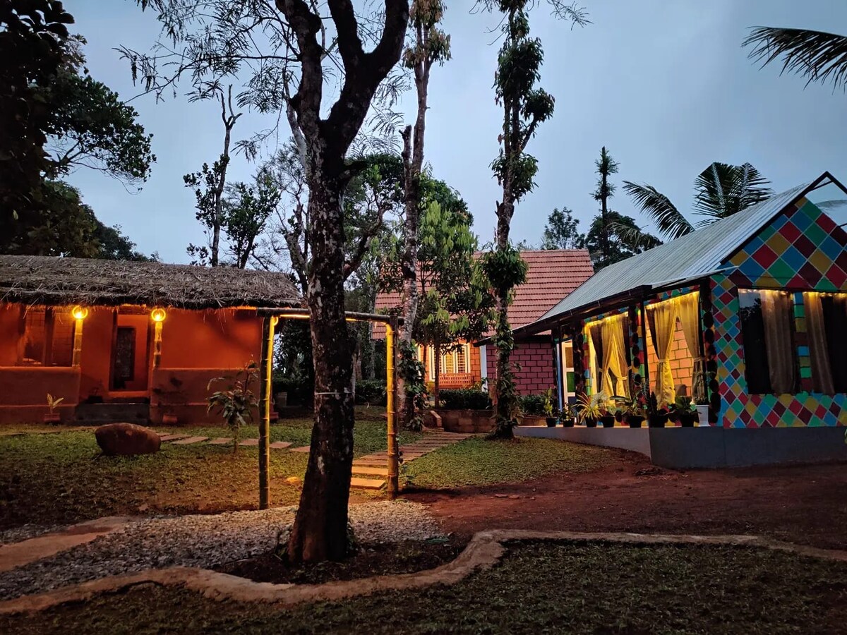 A serene outdoor scene features two distinct colorful structures surrounded by greenery. Soft lighting highlights the thatched-roof area on the left and the geometric-patterned house on the right. Towering trees frame the space, while a small gravel pathway guides the eye through the tranquil setting.