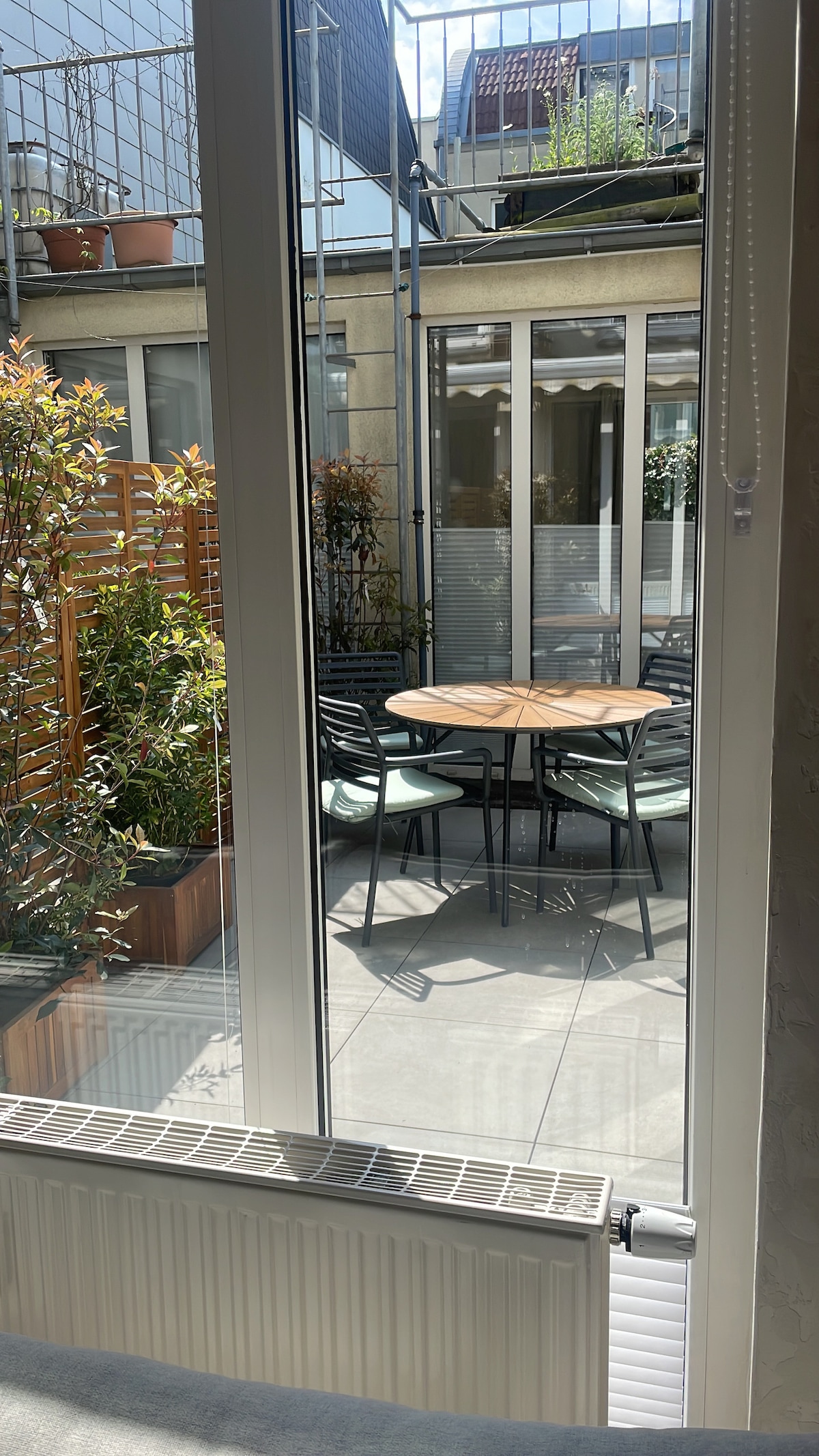 An outdoor terrace is visible through large glass doors, featuring a round wooden table surrounded by six black chairs. The space is enhanced by greenery in planters, with a concrete floor and soft natural light illuminating the area.