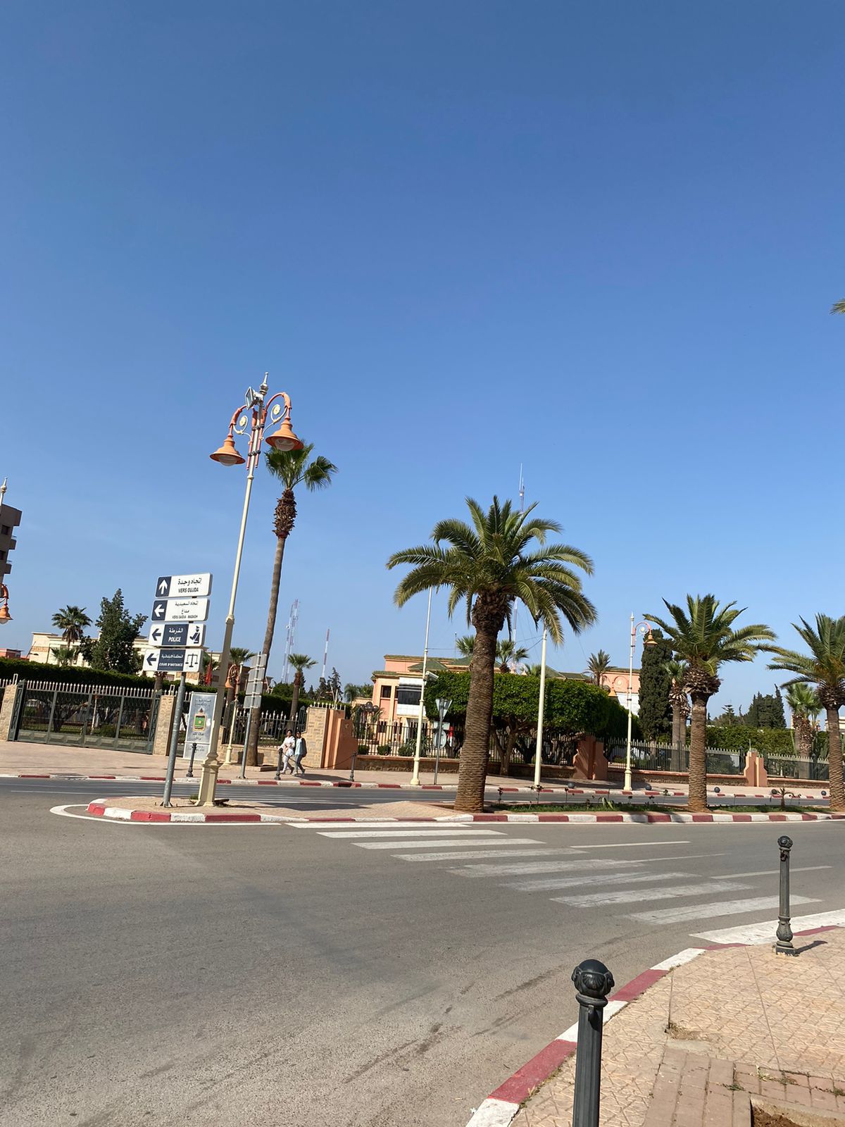 A vibrant street scene features a clear blue sky and palm trees along a wide roadway. Street lamps with decorative elements are visible, and pedestrians can be seen in the distance. The area appears well-maintained, with a blend of greenery and urban design.