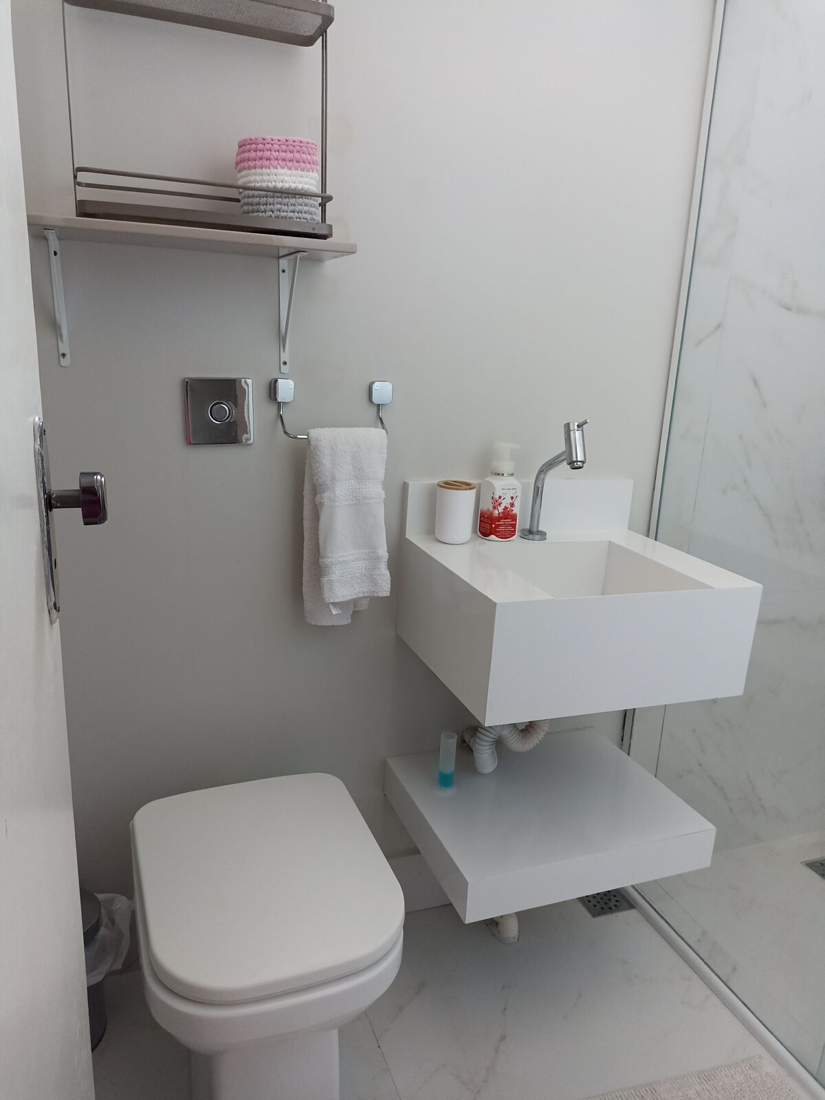 A modern bathroom features a sleek white toilet and a minimalist sink mounted on the wall. A small shelf holds neatly stacked toiletries, while a towel rack displays a crisp white towel. The glass shower enclosure is visible in the corner.