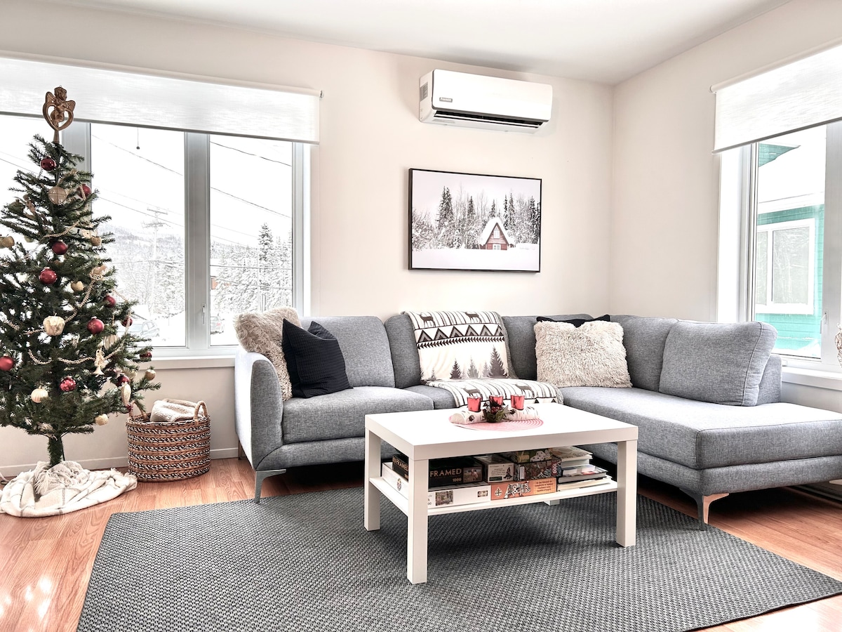 A cozy living area features a gray sectional sofa adorned with decorative pillows. A small coffee table holds books and a decorative tray. A Christmas tree stands in the corner, complemented by large windows allowing natural light to fill the space.