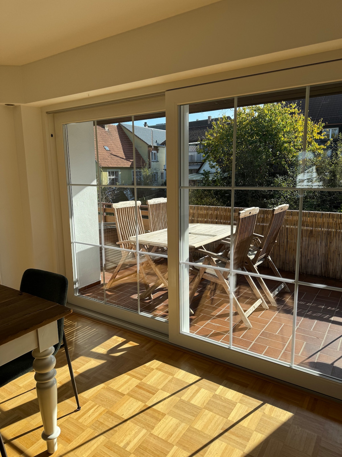 A bright dining area is shown through large glass doors that open to a private balcony. A wooden dining table is positioned next to the window, with several wooden chairs arranged around it. Sunlight filters in, highlighting the inviting atmosphere.