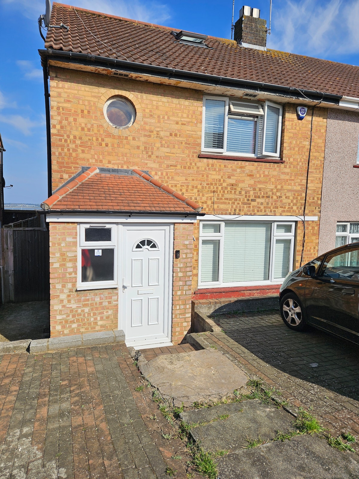 The image displays the exterior of a brick-clad residential property with a white front door. A circular window is visible above the entrance. A vehicle is parked in front of the house, and a well-maintained driveway can be seen, bordered by stone paving.
