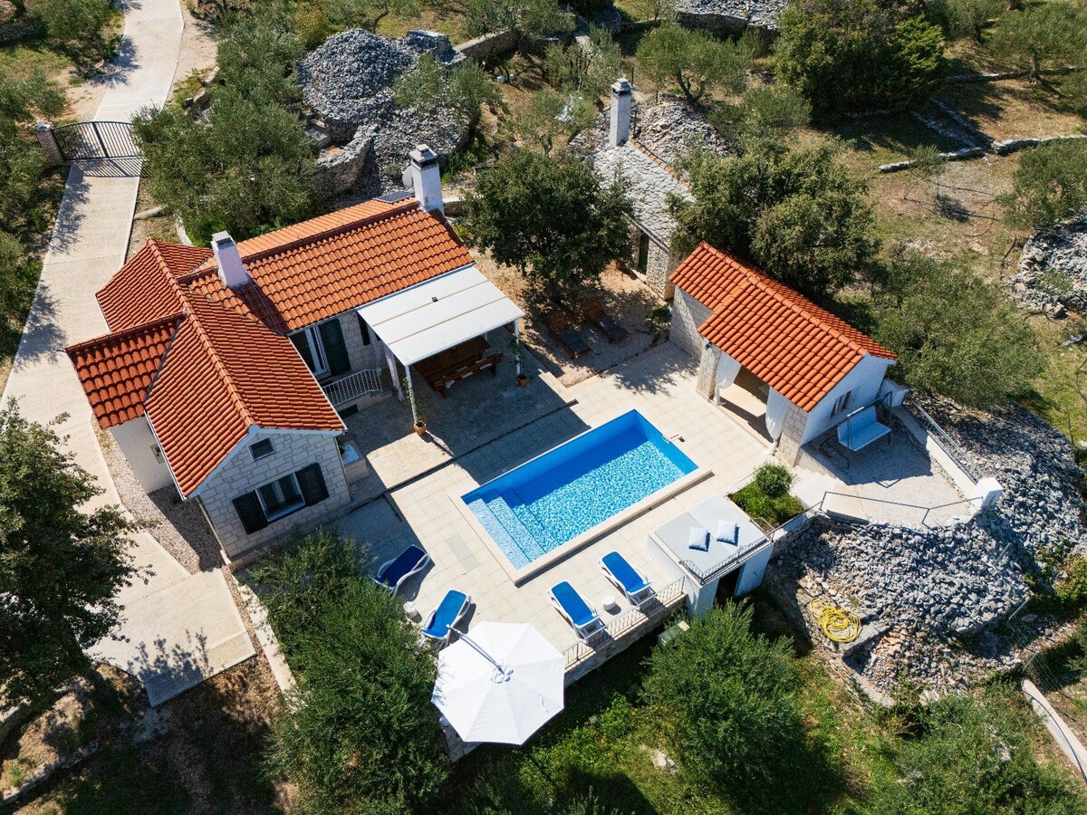 An aerial view of Casa Karlena showcases the modern country house surrounded by olive trees. The inviting pool occupies the center, complemented by lounge chairs. Side structures are visible, providing additional space. A shaded area is situated on the patio, enhancing outdoor relaxation options.