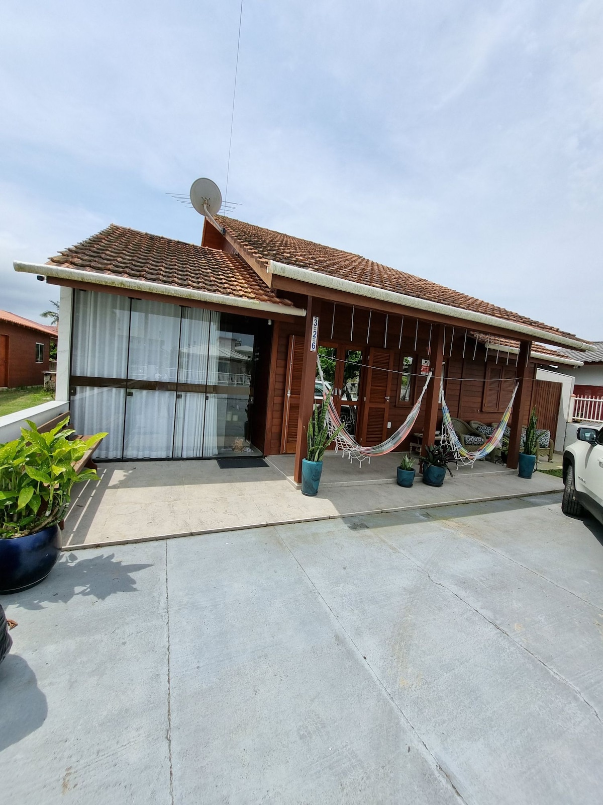 A charming wooden exterior features a covered porch decorated with hanging hammocks and various potted plants. Sunlight filters through sheer curtains, while a vehicle is parked nearby, complementing the welcoming entrance.
