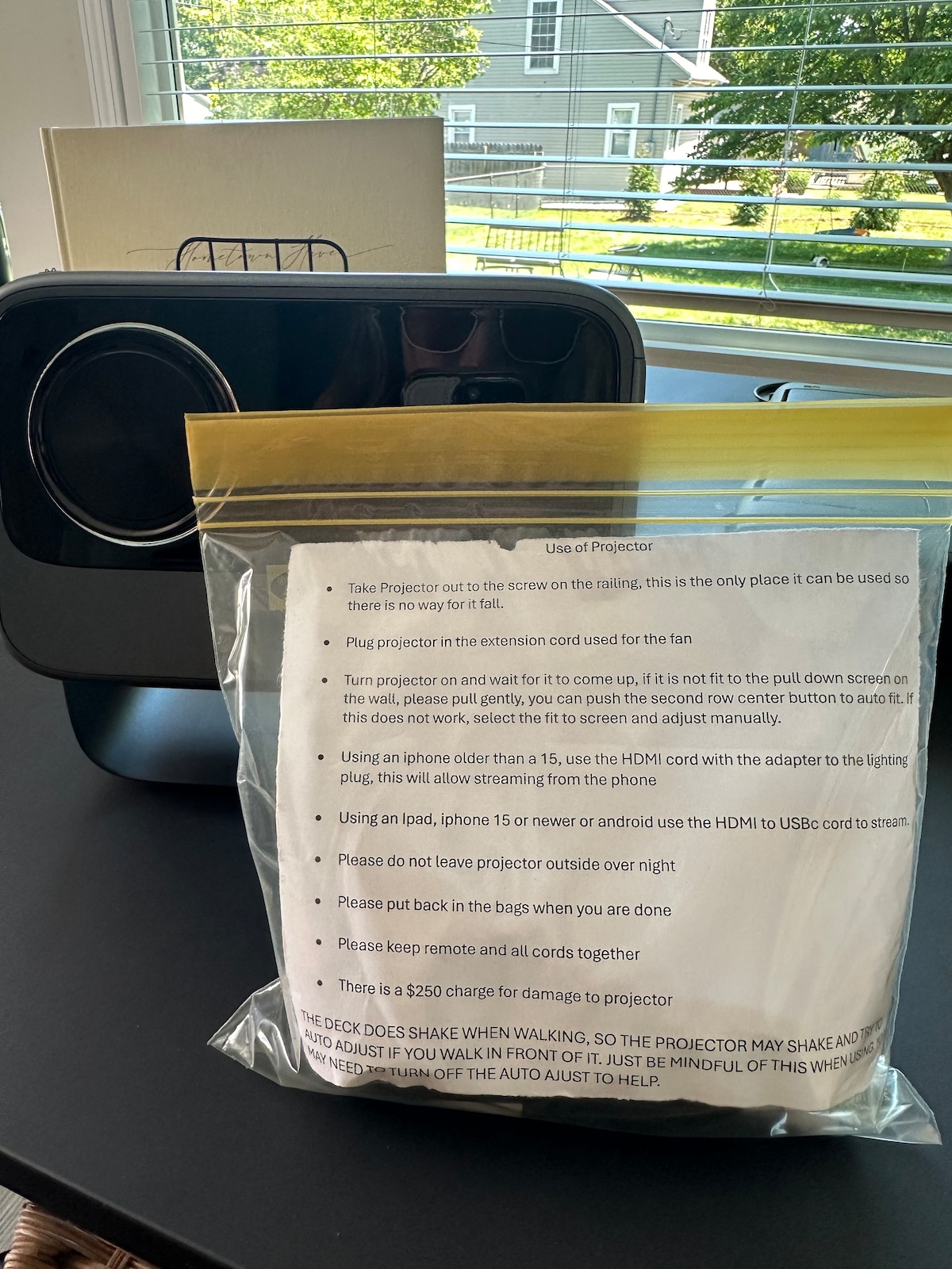 A projector is positioned on a stand, with a clear view of instructions in a plastic bag. Sunlight streams through the nearby window, creating a bright atmosphere. The area is neatly organized, enhancing accessibility for users looking to utilize the projector.