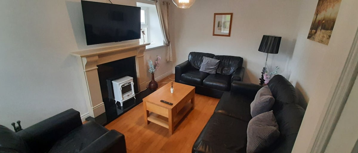 A welcoming living area features two dark leather sofas and a wooden coffee table, centered under a wall-mounted television. Soft lighting emanates from a nearby lamp, and a decorative fireplace adds a cozy touch. Complementary artwork decorates the walls.