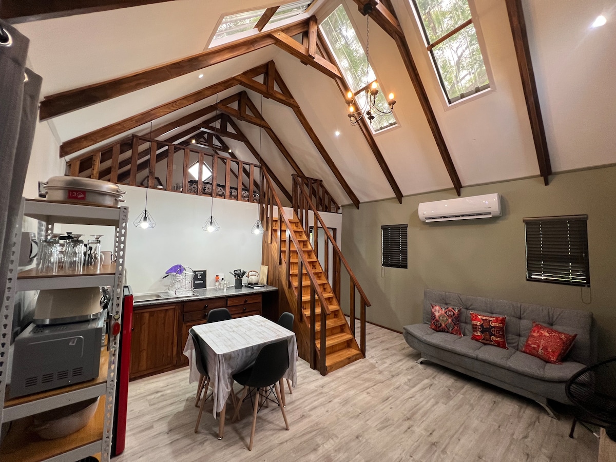 An open living space features high vaulted ceilings with exposed wooden beams. A staircase leads to a loft area. A dining table sits near the kitchen, and a comfortable sofa is arranged with decorative pillows. Natural light filters through skylights and windows.