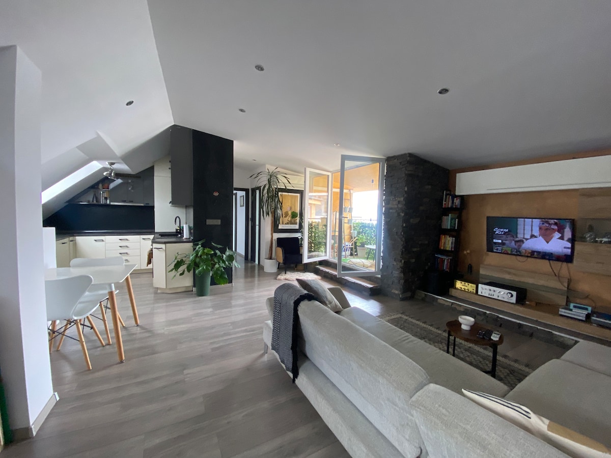 A bright living area features an open layout with modern furnishings. A cozy sofa is positioned near a wooden coffee table, while a television rests on a stone accent wall. Large glass doors open to a balcony, enhancing the space with natural light.