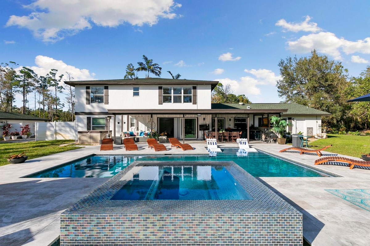 A spacious outdoor area features a modern pool surrounded by lounge chairs and a hot tub. The villa's two-story structure is visible in the background, flanked by palm trees and a lush green lawn, inviting relaxation and enjoyment.