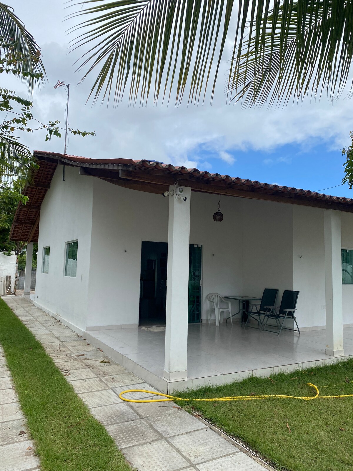 A single-story white house features a covered porch with a table and chairs, set against a backdrop of green grass. The sloped roof is decorated with clay tiles, and windows allow natural light to brighten the interior.